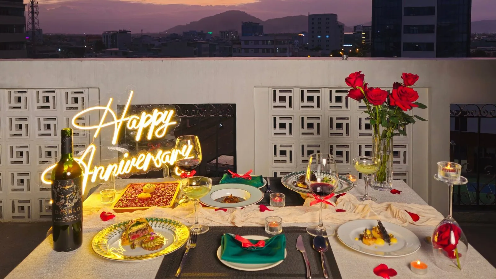 Bàn tiệc rooftop tại Le Bon Da Nang với lọ hoa hồng đỏ và biển neon Happy Anniversary